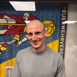 Ein lächelnder kahlköpfiger Mann in grauem Pullover vor einer bunten Wandgestaltung mit der Aufschrift Feuerwehrehrung