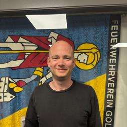 Ein lächelnder, kahler Mann in schwarzem Shirt steht vor einer bunten Feuerwehrvereinsfahne.